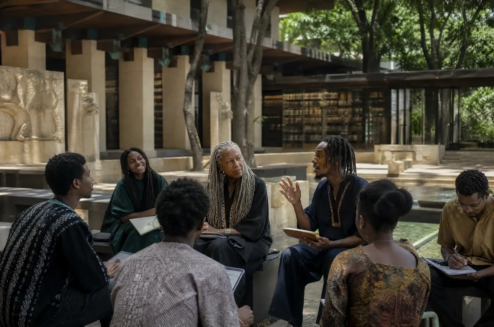 An outdoor seminar at the Royal University: a professor with grey locs listens to a student, a reading gallery of bound volumes behind smoked glass, a long water basin.